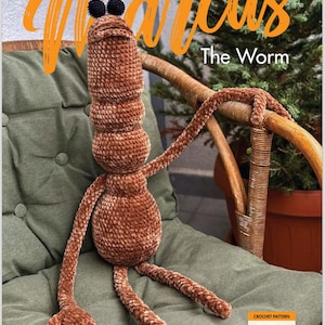 May include: A brown crocheted worm plush toy named "Marcus" with black pompom eyes and long, slender limbs. The worm is seated on a green cushion chair. The text "Marcus The Worm" is in orange script.