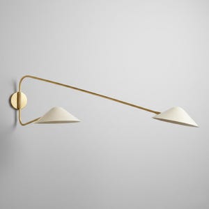May include: A modern wall sconce with a brass finish and two off-white cone-shaped shades. The fixture has a minimalist design, with a circular wall mount and a long, angled arm extending to the shades. The sconce is mounted on a light gray wall.