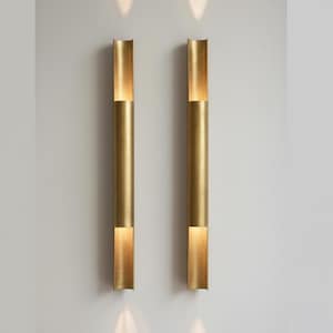 Peut inclure: Deux appliques murales verticales dorées de forme cylindrique. Chaque applique possède une ouverture rectangulaire en haut et en bas, émettant une lumière chaude. Les appliques sont fixées sur un mur de couleur neutre, créant une esthétique moderne. (Environ 100 cm de haut).