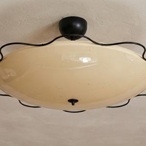 May include: A vintage ceiling light fitting featuring a cream-coloured, oval glass shade. The shade is encircled by a black, wavy metal frame. The fitting has a black base and a small, decorative black finial.