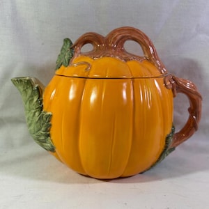 May include: A ceramic teapot shaped like a bright orange pumpkin with a glossy finish. The lid features a brown stem handle, while the spout is designed as a textured green leaf. A curved brown vine-like handle extends from the side of the body.