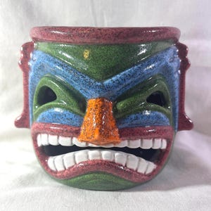 May include: A ceramic tiki mug in the shape of a tribal mask. The mug features a color palette of green, blue, red, and orange. The mask has a wide open mouth with white teeth, and eye holes. The mug is likely used for serving tropical drinks.