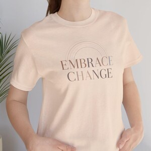 Op de afbeelding: Een licht perzikkleurig T-shirt met de tekst "EMBRACE CHANGE" in een bruin lettertype. De tekst is gebogen boven een halve cirkel. Het shirt heeft korte mouwen en een ronde hals.