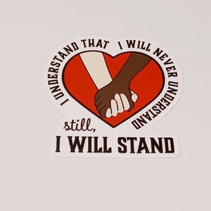 May include: A red heart with two hands holding each other, one white and one black. The text around the heart reads "I understand that I will never understand, still, I will stand".