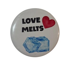 May include: White button with the words "LOVE MELTS" in black, a red heart, and two blue ice cubes. The button is round and has a glossy finish. The design is simple and graphic.