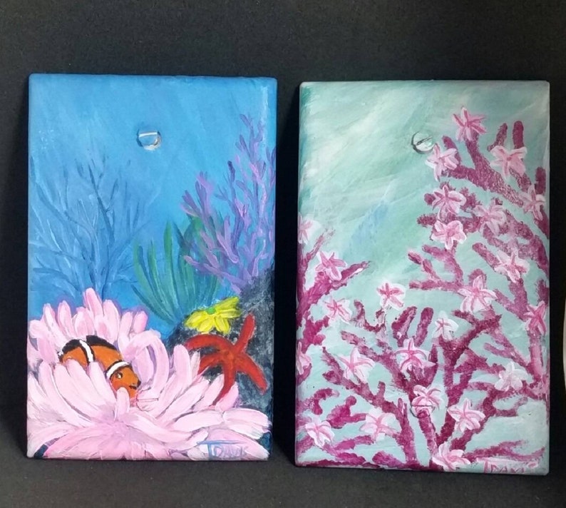 May include: Two small paintings on canvas depicting underwater scenes. The left painting shows a clownfish in a pink anemone with coral and a starfish. The right painting shows a pink coral reef with white flowers.