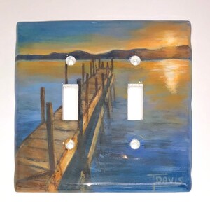 May include: A square light switch cover featuring a hand-painted landscape of a wooden pier extending into a body of water at sunset. The artwork includes shades of blue, orange, and brown. The cover has two switch openings and mounting holes.