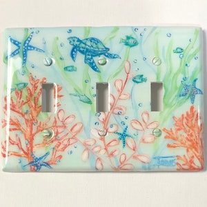 May include: A light blue, white, and green ceramic light switch cover with a sea life design. The design features a sea turtle, starfish, coral, and fish.