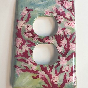 Ocean theme hand-painted on a vertical outlet receptacle wall plate cover with pink blooming coral and light grey color fish set against a blended light green color background. Shows a shiny reflective lighting spot indicating the artwork is protected with a resin glaze to ensure durability and give a glass like finish. Signed by Artist “TDavis” in the bottom right corner.