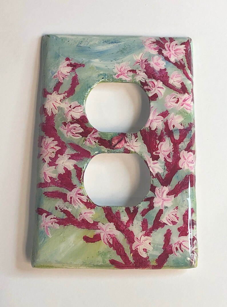 Ocean theme hand-painted on a vertical outlet receptacle wall plate cover with pink blooming coral and light grey color fish set against a blended light green color background. Shows a shiny reflective lighting spot indicating the artwork is protected with a resin glaze to ensure durability and give a glass like finish. Signed by Artist “TDavis” in the bottom right corner.
