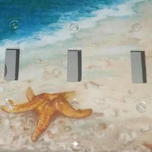 May include: A light switch cover with a beach scene painted on it. The scene includes a blue ocean, white sand, and a starfish. There are three light switch openings on the cover.