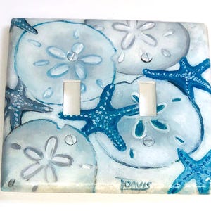 May include: A decorative light switch cover featuring a hand-painted design of sand dollars and starfish in shades of blue and white. The cover has two switch openings and is signed "J DAVIS" in the bottom right corner.