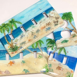 May include: Three painted light switch covers with a beach scene featuring palm trees, blue water, and sea turtles. The covers are painted in shades of blue, green, and brown. The text "Travis" is visible on one of the covers.