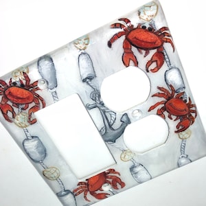 May include: A white light switch and outlet cover with a nautical theme. The cover is decorated with painted images of red crabs, anchors, and rope.
