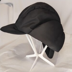 May include: A black winter hat with a visor and ear flaps. The hat is made of a smooth, padded material and is displayed on a white stand. The hat's design suggests warmth and protection from the cold.