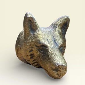 May include: A bronze-colored sculpture of a fox head, with detailed fur texture and pointed ears. The fox head is angled slightly to the side, showcasing its snout and facial features. The sculpture is likely a decorative item.