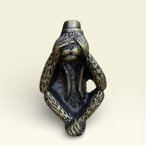 Op de afbeelding: Een bronzen aapfiguur in zittende positie, die zijn ogen met zijn handen bedekt. De sculptuur heeft een getextureerd oppervlak met gouden accenten en een kleine hoed. De gelaatstrekken van de aap zijn gedetailleerd.