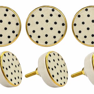 May include: A set of six round cabinet knobs featuring a cream-colored surface with a black polka dot pattern. Each knob is finished with a metallic gold-toned rim and includes a threaded metal screw extending from the back for installation.