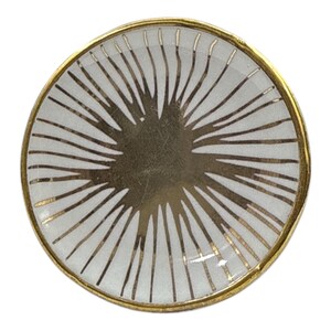 May include: A round decorative plate with a white background and gold radial lines. The plate has a gold rim. The design resembles a sunburst. The plate is suitable for decorative purposes. The diameter is approximately 15 cm.