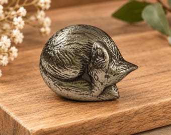 May include: A small, metallic, dark gray figurine of a curled-up, sleeping fox. The surface of the fox sculpture features a textured, cast-metal finish that highlights the shape of its body, ears, and tail tucked closely against its side.