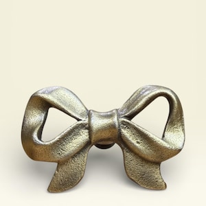 Pode incluir: Um puxador de armário em forma de laço dourado. O puxador tem uma superfície texturizada e uma pequena base redonda para montagem. O design do laço apresenta dobras e curvas detalhadas, dando-lhe uma aparência realista. A ferragem é provavelmente de metal.