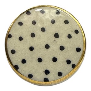 May include: A circular button featuring a cream-coloured surface adorned with black polka dots. The button is edged with a thin, gold-coloured metal. Ideal for sewing or craft projects, this button has a classic design.