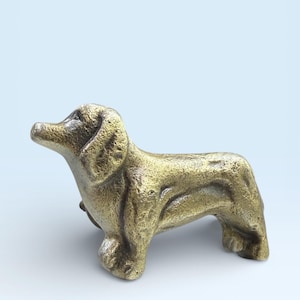 Puede incluir: Una figura de perro salchicha de color bronce. La escultura tiene una superficie texturizada y está representada en posición de pie, con la cabeza ligeramente levantada. Los rasgos del perro son detallados y el diseño general es realista.