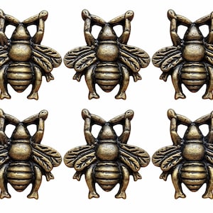 Peut inclure: Un ensemble de six boutons ou ornements décoratifs en métal en forme d'abeille. Chaque pièce présente une finition texturée couleur bronze antique avec des ailes détaillées, un corps segmenté et des pattes en relief, disposées en deux rangées de trois.