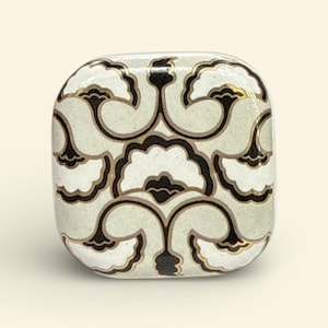 Könnte beinhalten: Quadratische dekorative Box mit einem sich wiederholenden Blumenmuster in Weiß, Schwarz und Gold auf hellgrünem Hintergrund. Die Box hat abgerundete Kanten und ein glänzendes Finish.