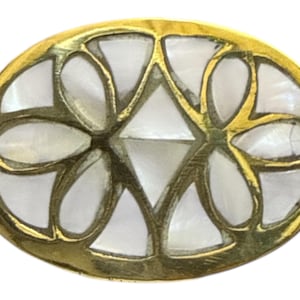 Puede incluir: Joya ovalada de metal dorado con diseño floral. El centro presenta una incrustación blanca en forma de diamante, rodeada de formas de pétalos. El borde exterior es de color dorado pulido.