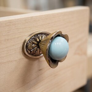 Peut inclure: Un bouton de tiroir composé d'un centre sphérique bleu clair lisse inséré dans un support en métal couleur laiton en forme de fleur. La plaque de base est ovale avec un motif floral en relief détaillé, fixée sur une surface en bois clair.