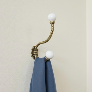 Op de afbeelding: Een decoratieve wandhaak met een messing afwerking en witte keramische balaccenten. De haak heeft een gedraaid touwontwerp en houdt een blauwe doek vast. De haak is bevestigd aan een crèmekleurige muur.