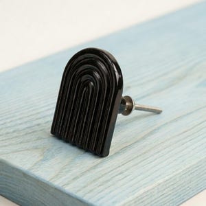 May include: A black cabinet knob featuring a raised, arched rainbow design with concentric semi-circular grooves. The hardware includes a silver-toned metal bolt extending from the back of the glossy, curved, arch-shaped decorative pull.