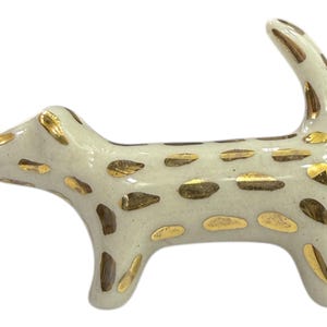 Op de afbeelding: Een keramieken beeldje van een gestileerde hond in staande houding. De hond is gebroken wit met goudkleurige vlekken en accenten. De staart is omhoog gebogen. Een decoratief item.