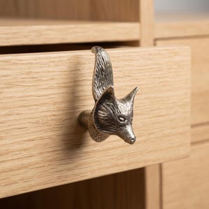 May include: A silver-toned metal drawer knob shaped like a fox head with an upright tail. The fox features detailed fur texture and pointed ears, mounted on a light-coloured wooden drawer front with a visible horizontal wood grain pattern.