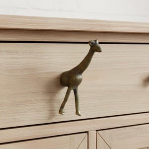 Peut inclure: Poignée de tiroir en forme de girafe de couleur bronze, fixée sur une armoire en bois clair. La girafe est représentée debout, la tête et le cou tendus vers le haut. L'armoire présente un fini grain de bois naturel.