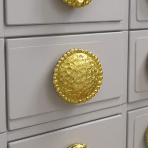 Op de afbeelding: Close-up van een lichtgrijze kast met een goudkleurige, getextureerde knop. De knop is rond met een getextureerd oppervlak en een kralenrand. De kast heeft een rechthoekige vorm.