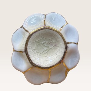Puede incluir: Un pequeño plato de cerámica circular con borde festoneado, que presenta un acabado blanco brillante en los pétalos exteriores y una zona central hundida con un esmalte de color crema agrietado. Los segmentos están unidos por finas líneas doradas.