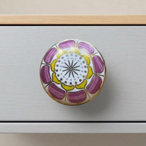 Op de afbeelding: Een ronde meubelknop met een decoratief bloemmotief met paarse en gele bloemblaadjes rondom een witte cirkel met een zwart sterontwerp en goudkleurige metalen randen, gemonteerd op een lichtgrijze houten ladefront.