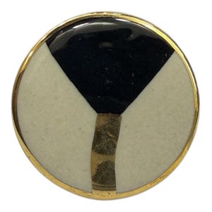 May include: A circular pin with a gold-coloured border. The pin's face has a black triangle at the top, with two off-white sections either side. A gold-coloured vertical accent runs down the centre.