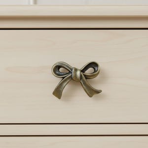 Może przedstawiać: Brązowy uchwyt do szuflady w kształcie kokardy zamontowany na jasnej drewnianej szufladzie. Kokarda ma szczegółowe krzywizny i estetykę vintage, dodając meblom dekoracyjnego charakteru. Szuflada jest wykonana z jasnego drewna.