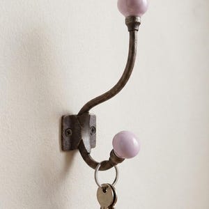 May include: A wall-mounted metal hook with a dark, weathered finish featuring two curved arms. Each arm ends in a smooth, round, light lavender ceramic knob. A silver-toned metal keyring with two keys hangs from the lower hook arm.