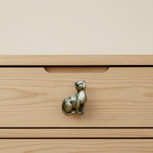 以下が含まれることがあります： 真鍮製の猫型引き出しつまみが明るい木製の引き出しに取り付けられています。猫は座っており、尻尾が丸まっています。引き出しには開けるための水平な溝があります。背景はニュートラルなベージュです。