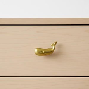 Puede incluir: Un tirador de cajón dorado con forma de ballena. La ferretería decorativa está montada en un cajón de madera de color claro. El tirador de ballena tiene un acabado liso y está diseñado para agarrarse fácilmente.