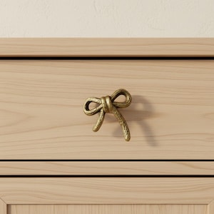 May include: A gold-colored bow-shaped drawer pull is mounted on a light-colored wooden drawer. The bow handle adds a decorative touch to the drawer's design. The drawer is made of light-colored wood.