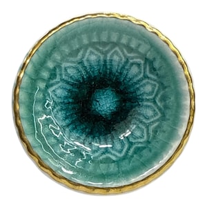 Op de afbeelding: Een ronde, turquoise keramische schaal met een goudkleurige rand. De schaal heeft een getextureerd, gecraqueleerd oppervlak met een donkerder turquoise centrum. Het ontwerp heeft een bloemenpatroon.