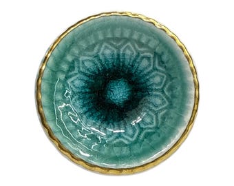 May include: A round, teal-colored ceramic dish with a gold-colored rim. The dish has a textured, crackled surface with a darker teal center. The design has a floral pattern.