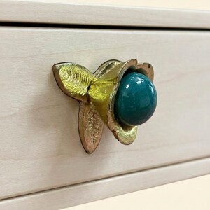 Peut inclure: Un bouton de tiroir composé d'une base en laiton doré en forme de fleur avec des feuilles texturées et une perle ronde, lisse et bleu sarcelle foncé, fixée au centre sur une façade de tiroir en bois de couleur claire.