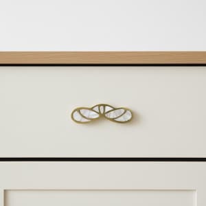 Könnte beinhalten: Dekorative Schrankgriff mit Messing-Finish und eingelegtem Perlmutt-Design. Der Griff hat die Form einer stilisierten Blume oder Schleife und ist an einer weißen Schranktür befestigt. Die Oberseite des Schranks ist helles Holz.