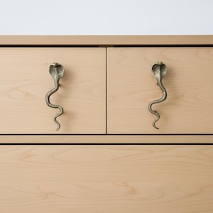 Könnte beinhalten: Zwei hellfarbene Holzschubladen mit messingfarbenen, kobraförmigen Griffen. Die Griffe sind mit einem Schlangenkopf und einem geschwungenen Körper versehen. Der Schrank hat ein schlichtes, modernes Design.